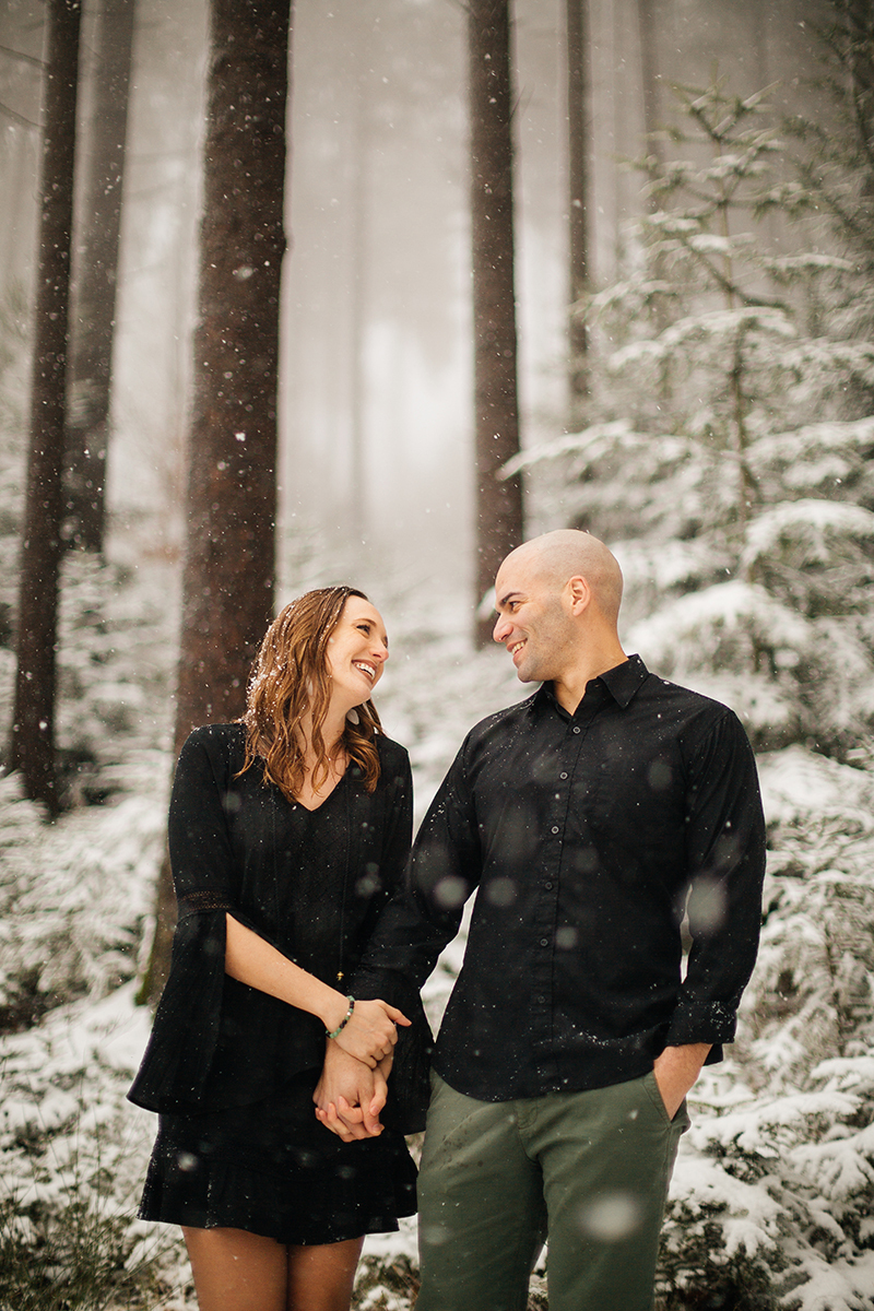 A couple stand side by side in a snow covered forest in Germany wearing a beautiful black dress and black button up shirt for a Black Forest engagement photography session