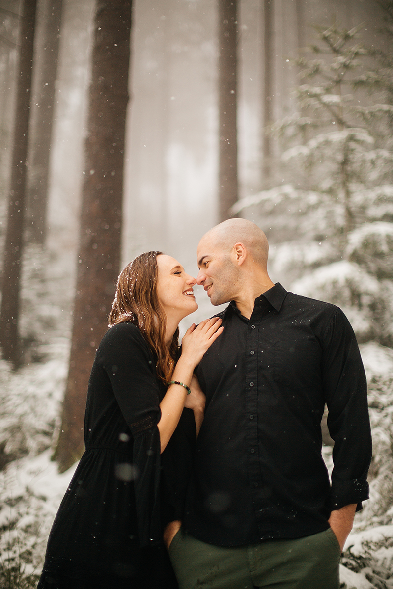 A couple stand side by side in a snow covered forest in Germany wearing a beautiful black dress and black button up shirt for a Black Forest engagement photography session