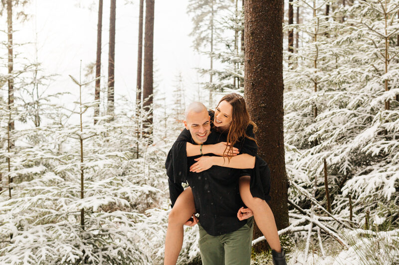 A couple embrace as he gives her a piggyback ride in a snow covered forest in Germany wearing a beautiful black dress and black button up shirt for a Black Forest engagement photography session
