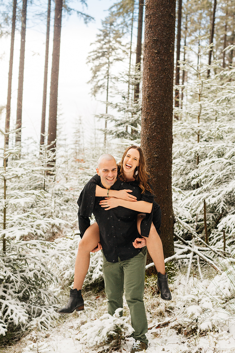 A couple embrace as he gives her a piggyback ride in a snow covered forest in Germany wearing a beautiful black dress and black button up shirt for a Black Forest engagement photography session