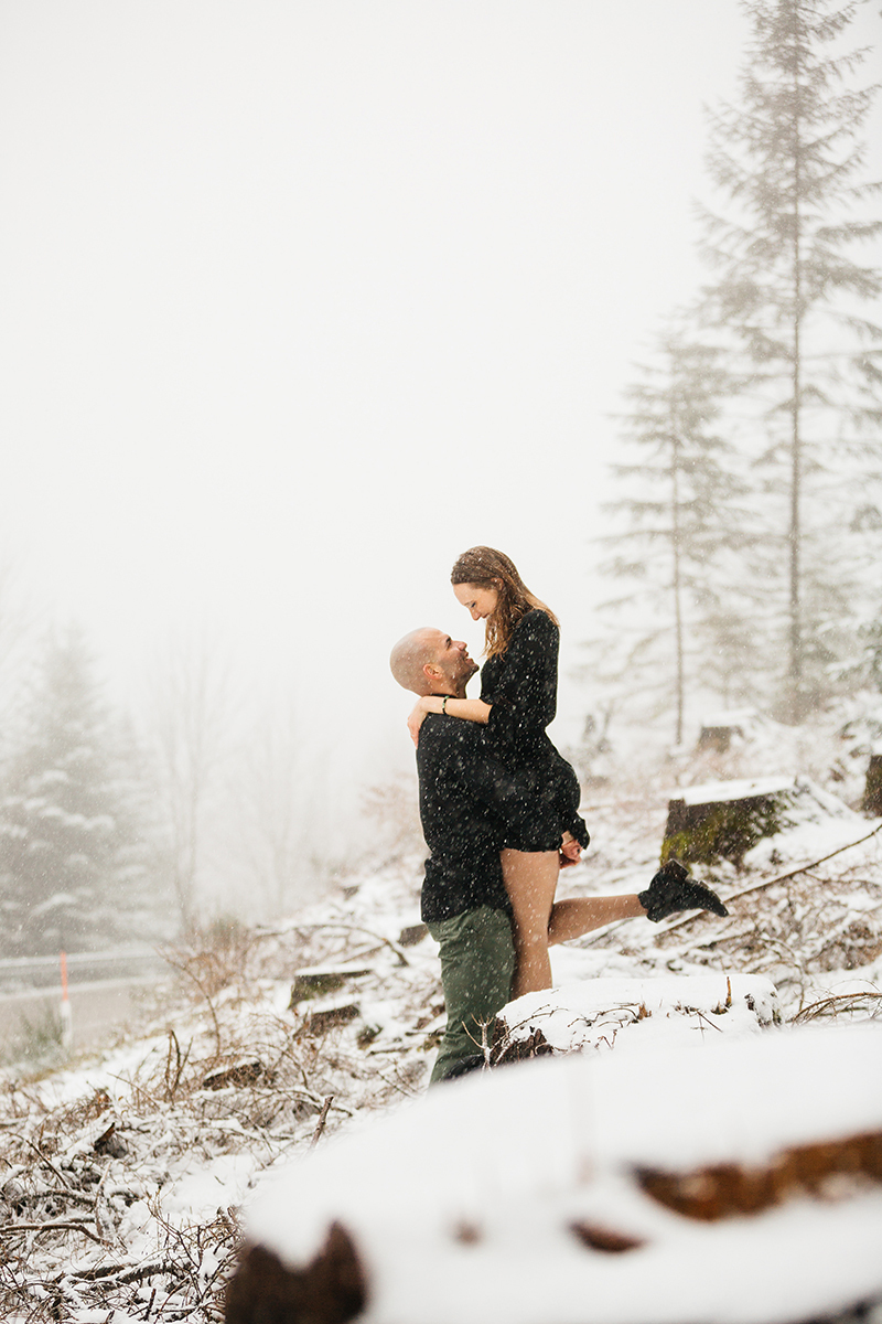 A couple embraces as he lifts her up in a snow covered forest in Germany wearing a beautiful black dress and black button up shirt for a Black Forest engagement photography session