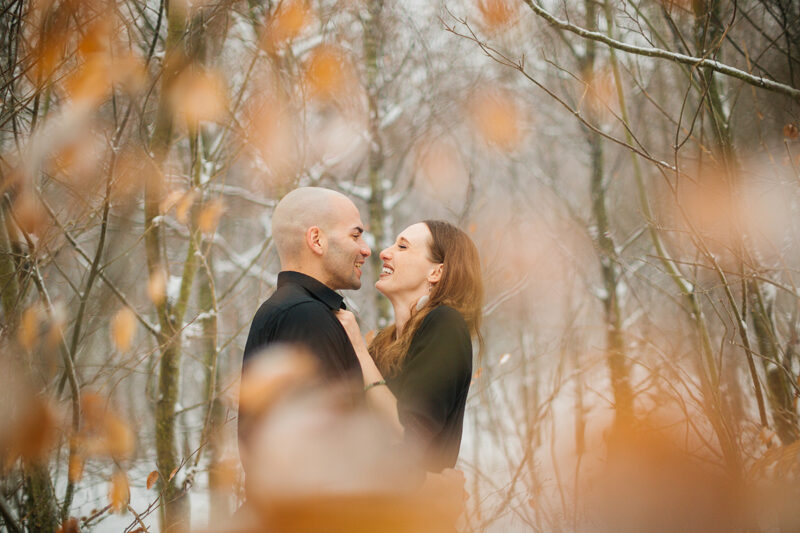 A couple embraces in a snow covered forest in Germany wearing a beautiful black dress and black button up shirt for a Black Forest engagement photography session