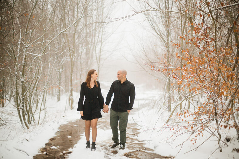 A couple holds hands as they walk in a snow covered forest in Germany wearing a beautiful black dress and black button up shirt for a Black Forest engagement photography session