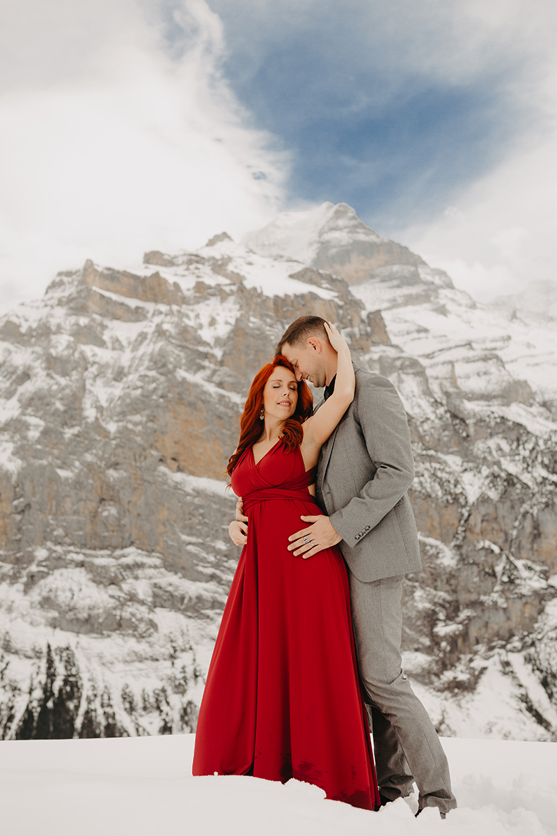 A couple embrace on a snow covered mountain in Switzerland wearing a beautiful red dress and gray suit for a Mürren couples photography session