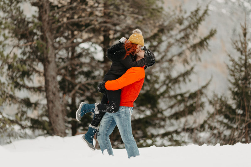 A couple embrace and kiss as he lifts her up in trees on a snow covered mountain in Switzerland wearing snow gear for a Mürren couples photography session