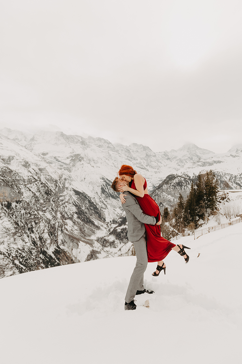 A couple embrace and kiss as he lifts her up on a snow covered mountain in Switzerland wearing a beautiful red dress and gray suit for a Mürren couples photography session