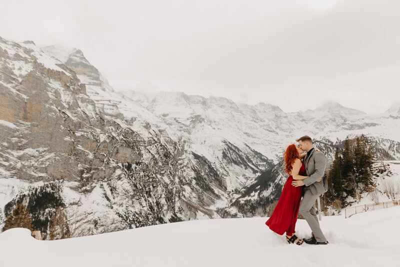 A couple embrace and kiss on a snow covered mountain in Switzerland wearing a beautiful red dress and gray suit for a Mürren couples photography session