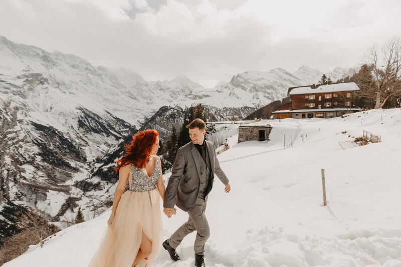 A couple holds hands walking up a snow covered mountain in Switzerland wearing a beautiful peach colored dress and gray suit for a Mürren couples photography session