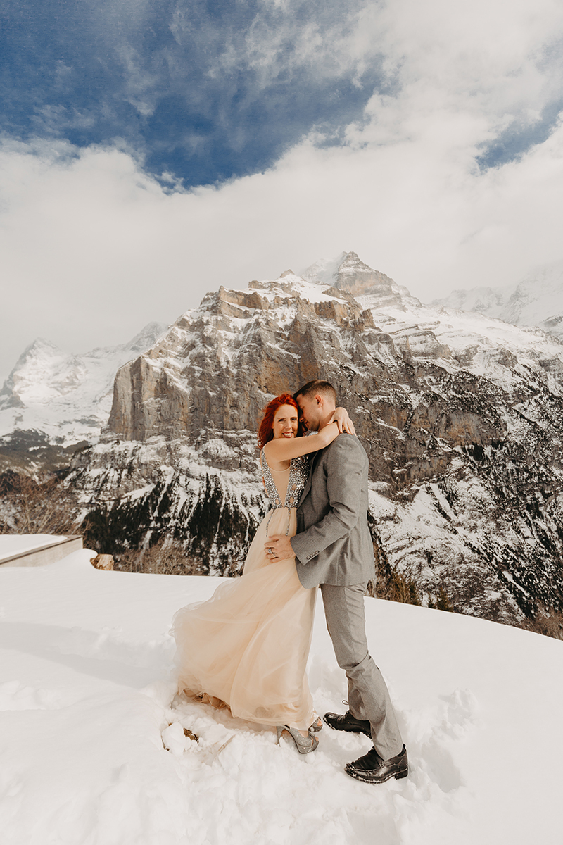 A couple embraces on a snow covered mountain in Switzerland wearing a beautiful peach colored dress and gray suit for a Mürren couples photography session
