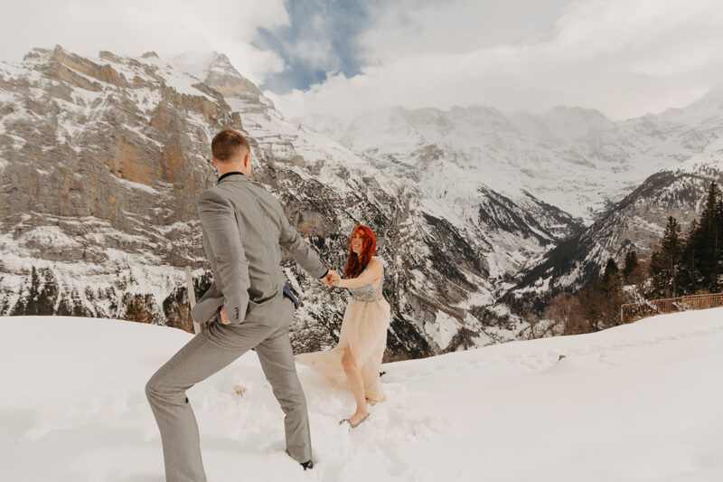 A couple holds hands walking up a snow covered mountain in Switzerland wearing a beautiful peach colored dress and gray suit for a Mürren couples photography session