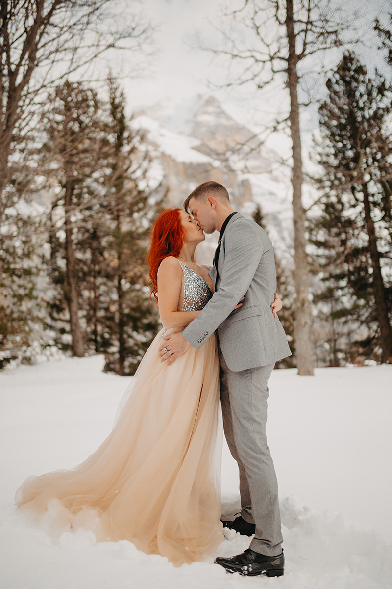 A couple embrace and kiss in trees on a snow covered mountain in Switzerland wearing a beautiful peach colored dress and gray suit for a Mürren couples photography session