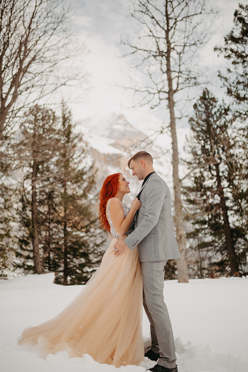 A couple embrace in trees on a snow covered mountain in Switzerland wearing a beautiful peach colored dress and gray suit for a Mürren couples photography session