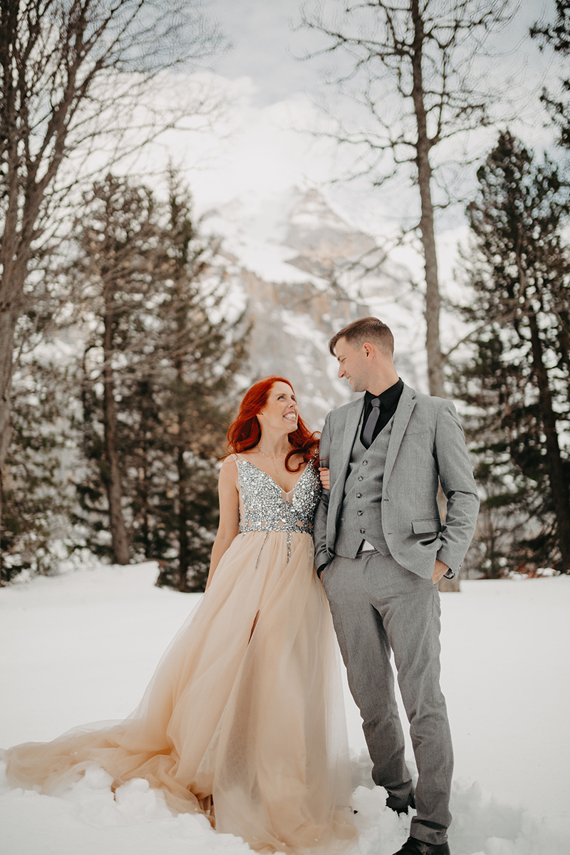 A couple walks side by side in trees on a snow covered mountain in Switzerland wearing a beautiful peach colored dress and gray suit for a Mürren couples photography session