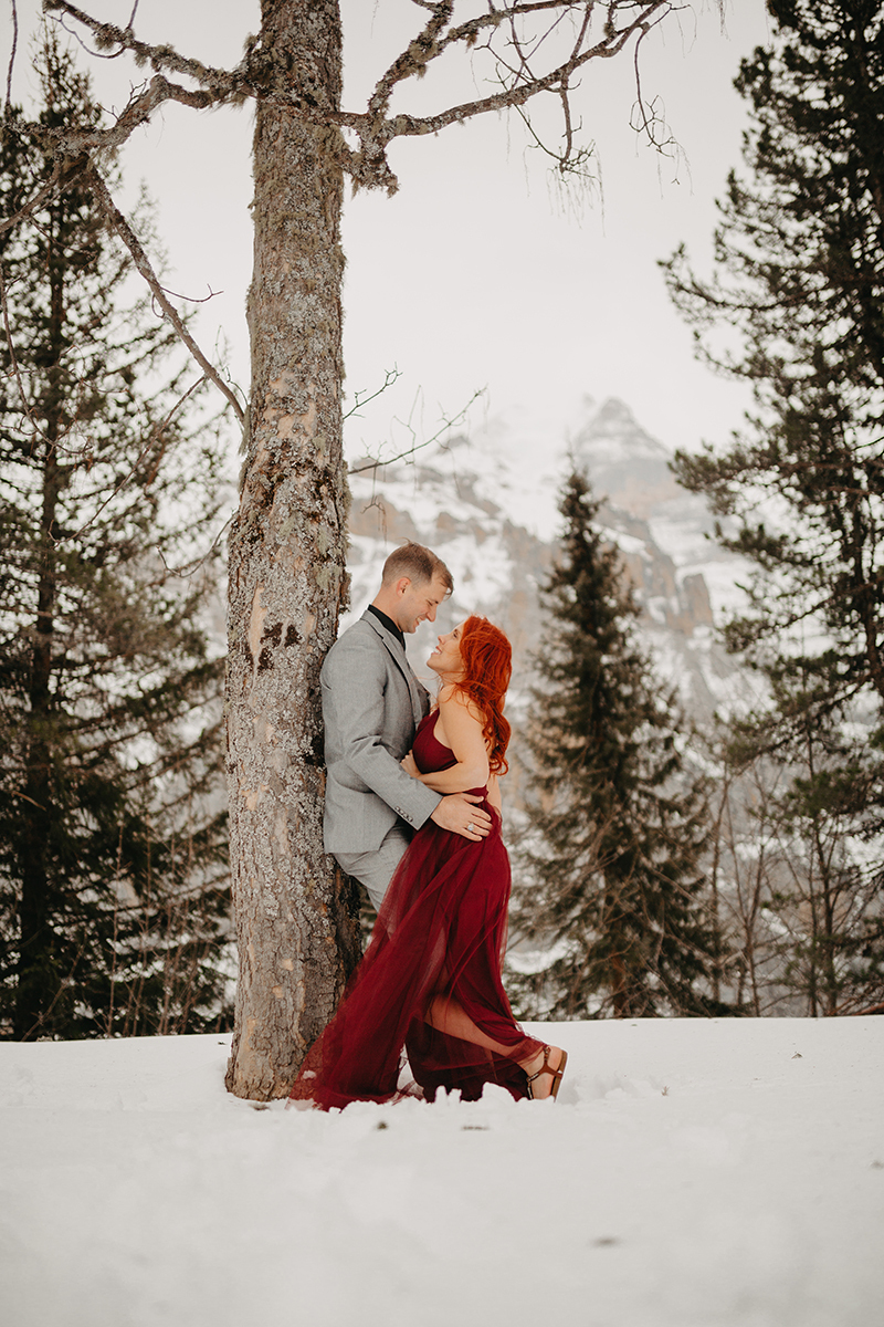 A couple embraces in trees on a snow covered mountain in Switzerland wearing a beautiful red dress and gray suit for a Mürren couples photography session
