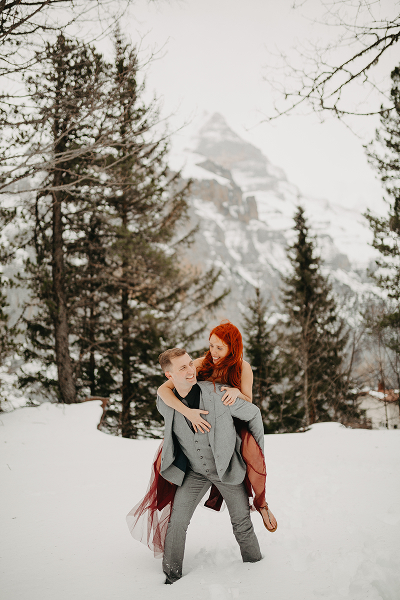 A couple walks as he gives her a piggyback ride in trees on a snow covered mountain in Switzerland wearing a beautiful red dress and gray suit for a Mürren couples photography session