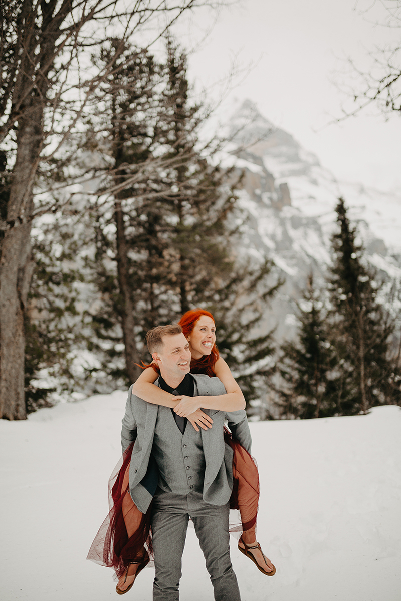 A couple walks as he gives her a piggyback ride in trees on a snow covered mountain in Switzerland wearing a beautiful red dress and gray suit for a Mürren couples photography session
