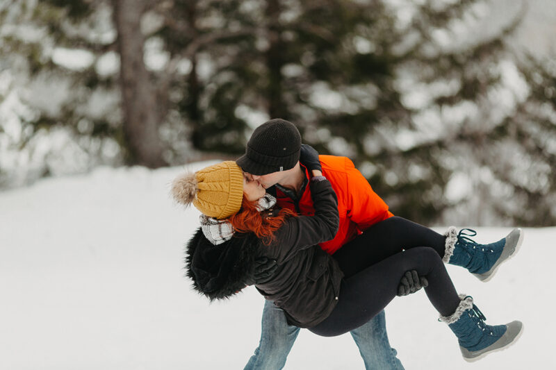 A couple embraces in trees on a snow covered mountain in Switzerland wearing snow gear for a Mürren couples photography session