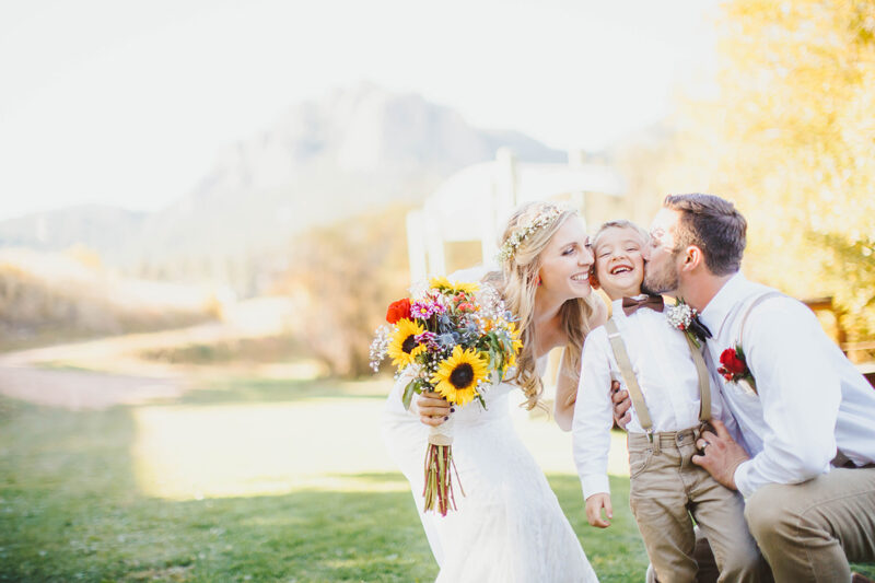 A bride and groom stand together kissing their son at sunset wearing a white dress and formal wear for this Lower Lake Ranch wedding photography session