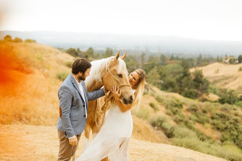 An engaged couple pet their horse on the hillside for this Granada Hills engagement photography session