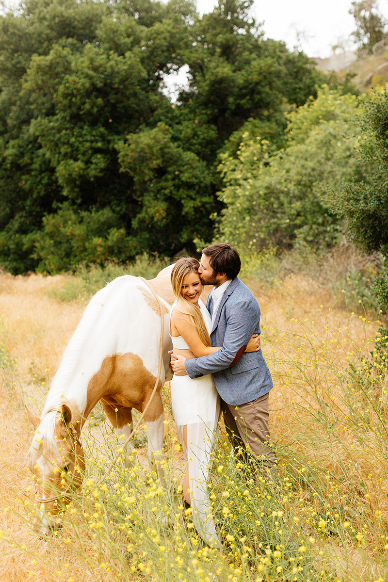 An engaged couple kiss holding each other with their horse on the hillside for this Granada Hills engagement photography session