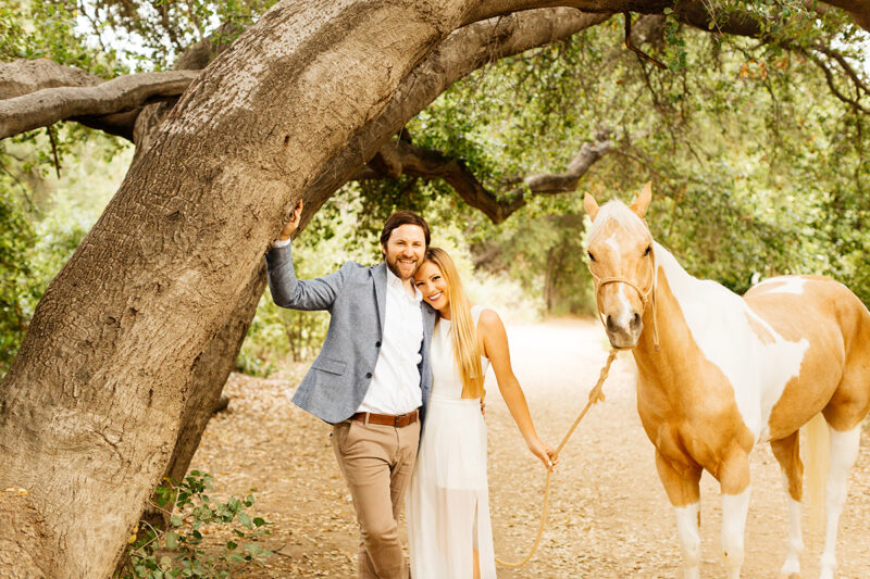 An engaged couple lean against a tree holding each other with their horse on a trail for this Granada Hills engagement photography session