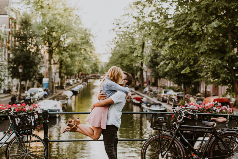 A couple hold one another close and kiss as he lifts her up on a bridge over a canal for this Amsterdam couples photography session