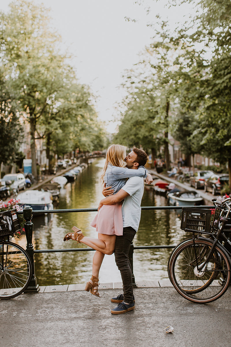 A couple hold one another close and kiss as he lifts her up on a bridge over a canal for this Amsterdam couples photography session