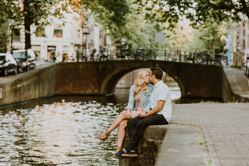 A couple sit together and kiss on the edge of a canal for this Amsterdam couples photography session