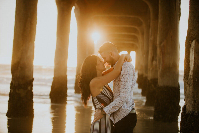 A couple hold each other close at sunset on the beach under a pier near the Santa Monica Pier for this Los Angeles engagement photography session