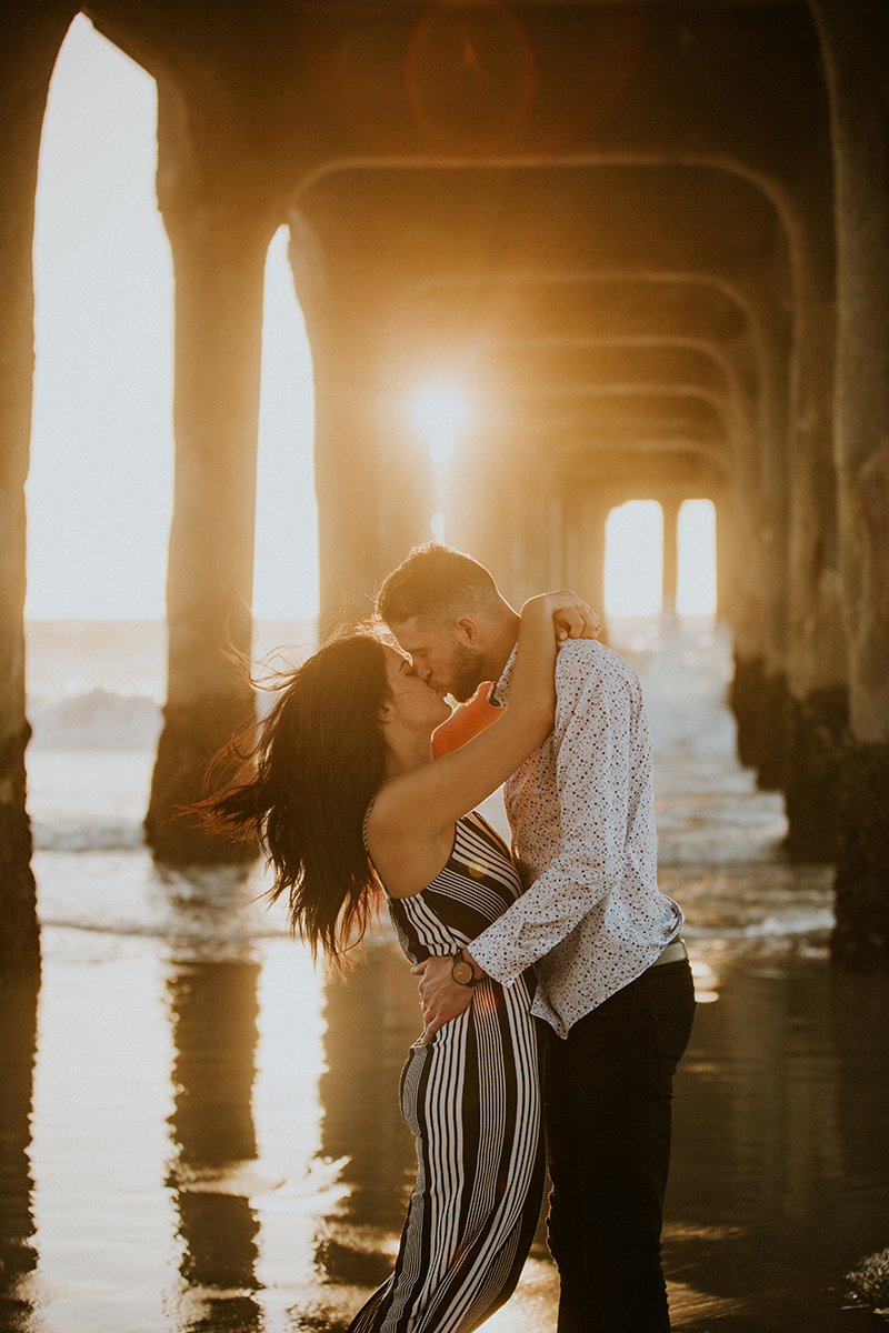 A couple kiss at sunset on the beach under a pier near the Santa Monica Pier for this Los Angeles engagement photography session