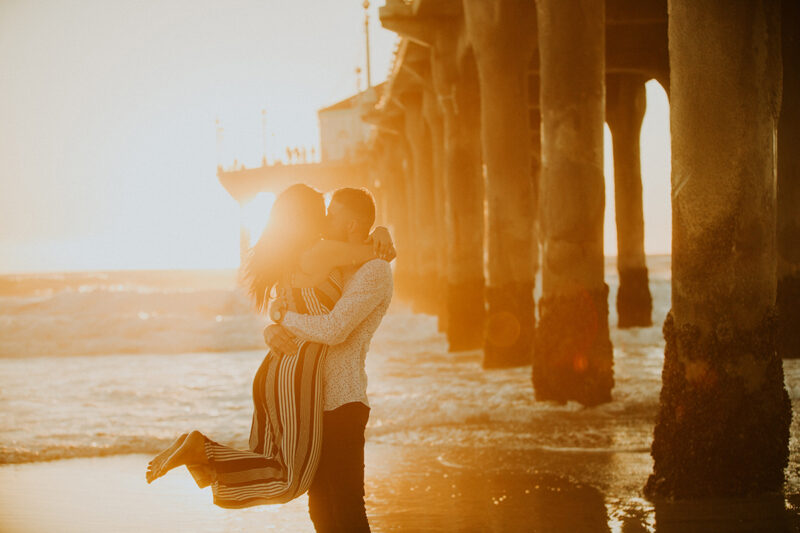 A couple kiss at sunset as he lifts her up on the beach near the Santa Monica Pier for this Los Angeles engagement photography session