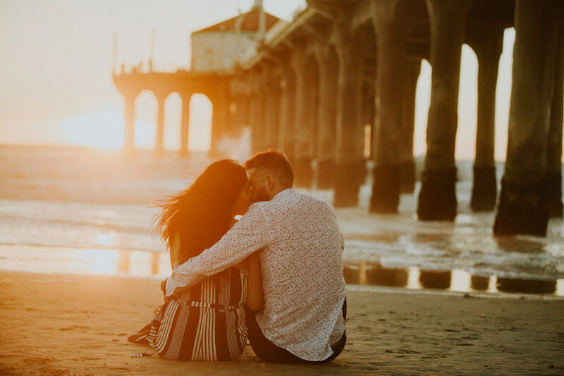 A couple sit together and kiss at sunset on the beach near the Santa Monica Pier for this Los Angeles engagement photography session