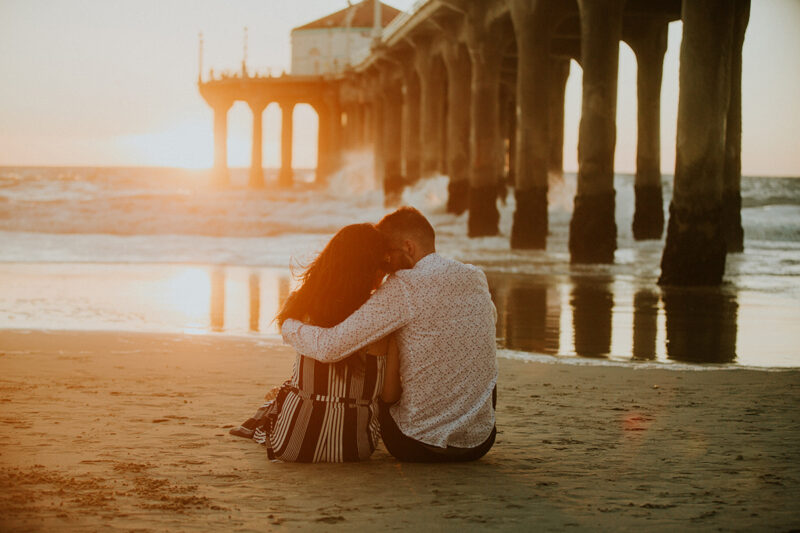 A couple sit together at sunset on the beach near the Santa Monica Pier for this Los Angeles engagement photography session