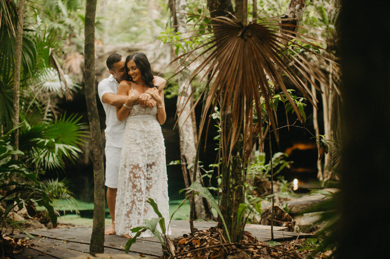 A couple hold one another smiling in front of a cenote in Mexico wearing a beautiful white dress and white shorts with a shirt for a Cenote Azul engagement photography session