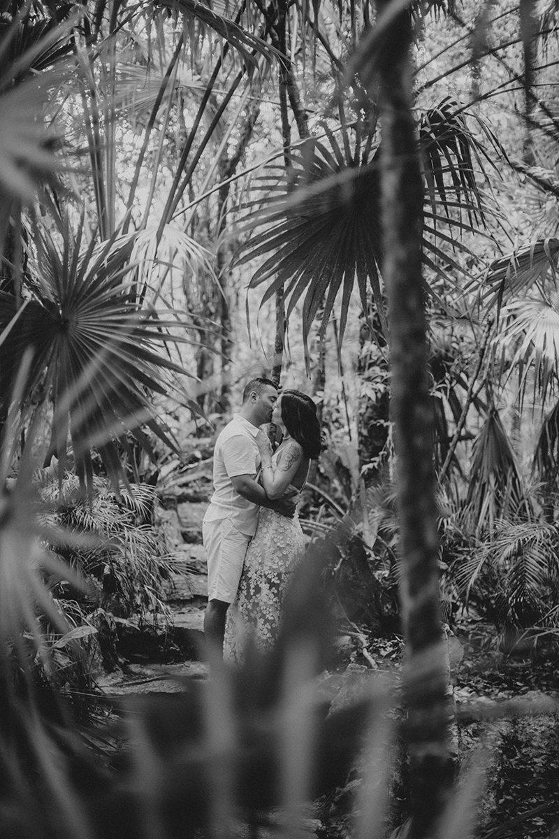 A couple hold one another kissing in front of a cenote in Mexico wearing a beautiful white dress and white shorts with a shirt for a Cenote Azul engagement photography session