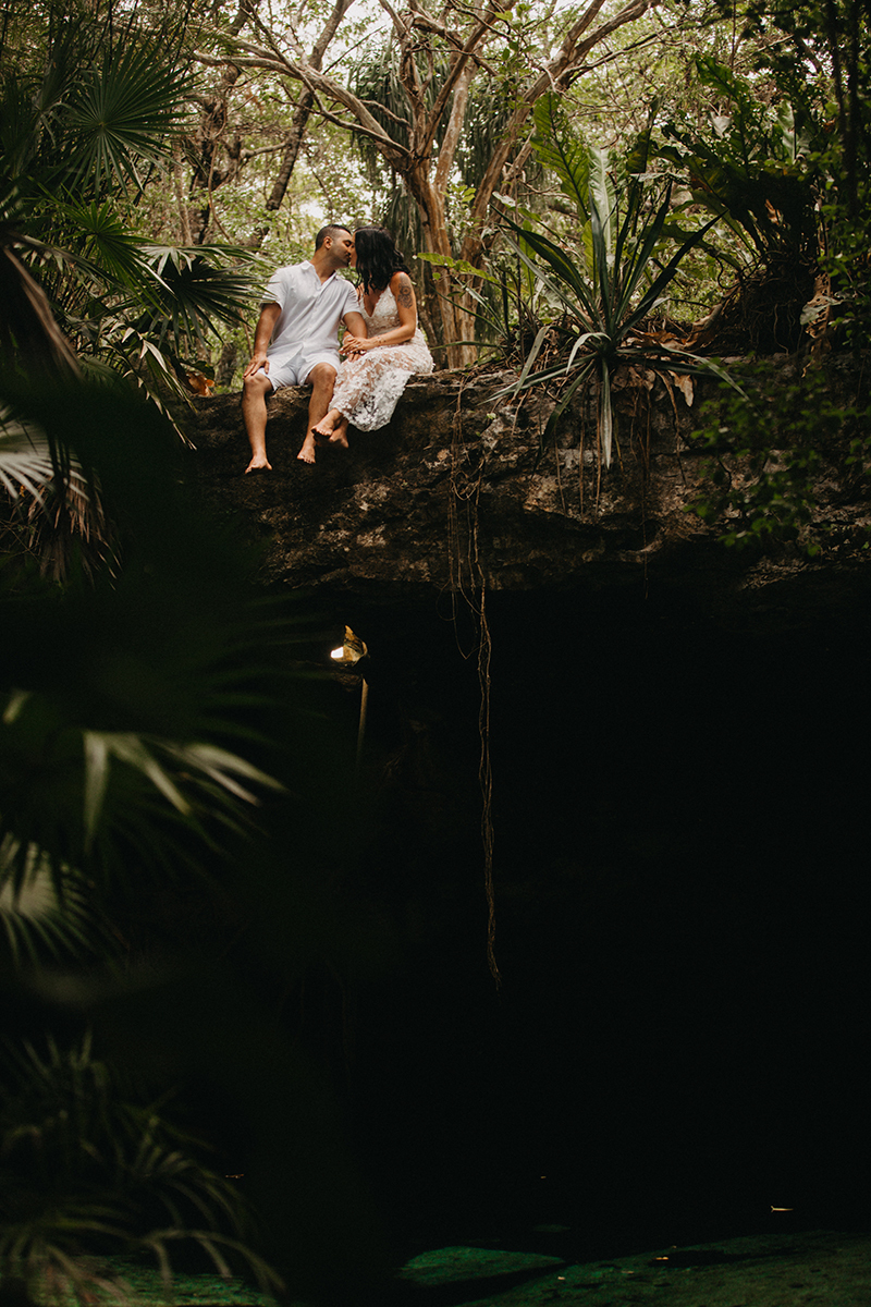 A couple sit next to each other kissing on top of a cenote in Mexico wearing a beautiful white dress and white shorts with a shirt for a Cenote Azul engagement photography session