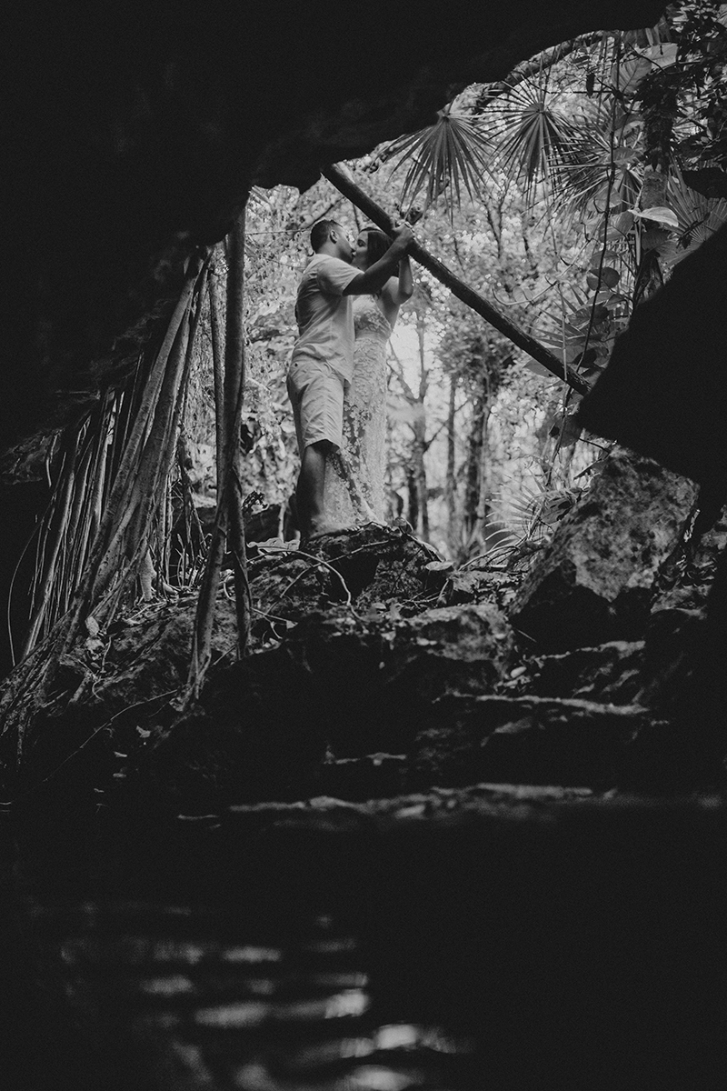 A couple together kissing at the edge of a cenote in Mexico wearing a beautiful white dress and white shorts with a shirt for a Cenote Azul engagement photography session