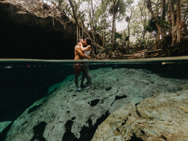A couple stand together kissing in the water of a cenote in Mexico wearing a white swimsuit and orange shorts for a Cenote Azul engagement photography session
