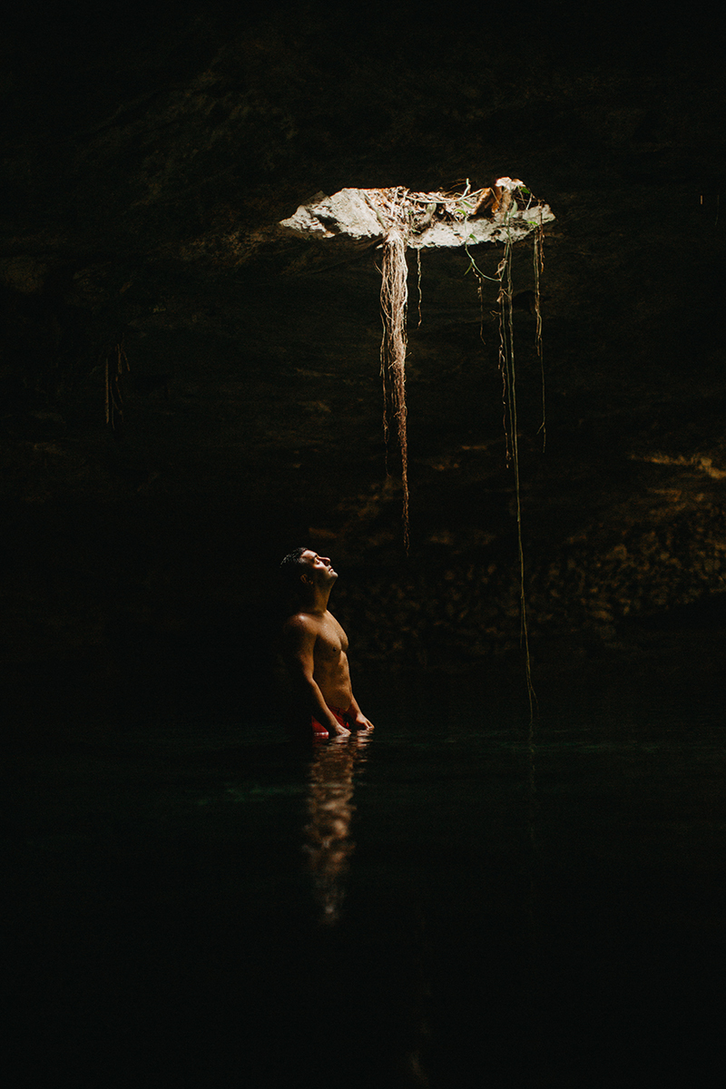 A handsome man poses in the water of a cenote in Mexico wearing orange shrts for a Cenote Azul engagement photography session