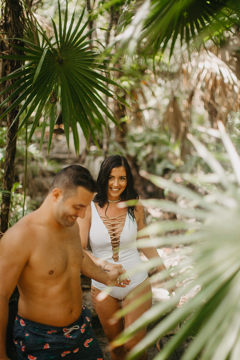 A couple walk together through the jungle near a cenote in Mexico wearing a white swimsuit and orange shorts for a Cenote Azul engagement photography session