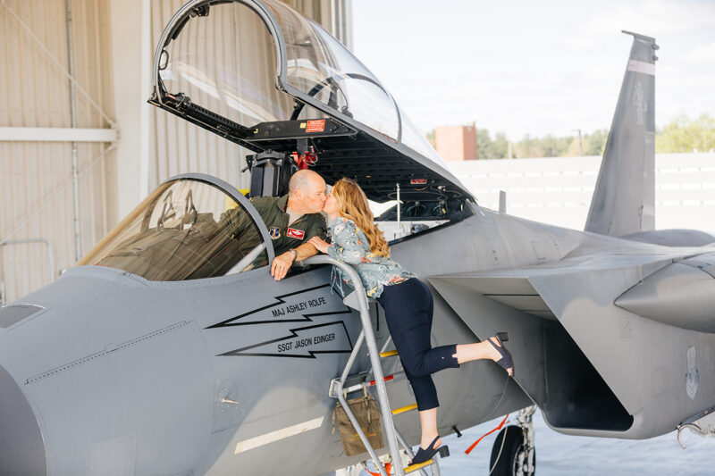A pilot sits in the cockpit of an F-15 with his wife on a ladder kissing him at Barnes Air National Guard Base wearing a flight suit and a coordinated outfit for these F-15 fighter pilot family photos