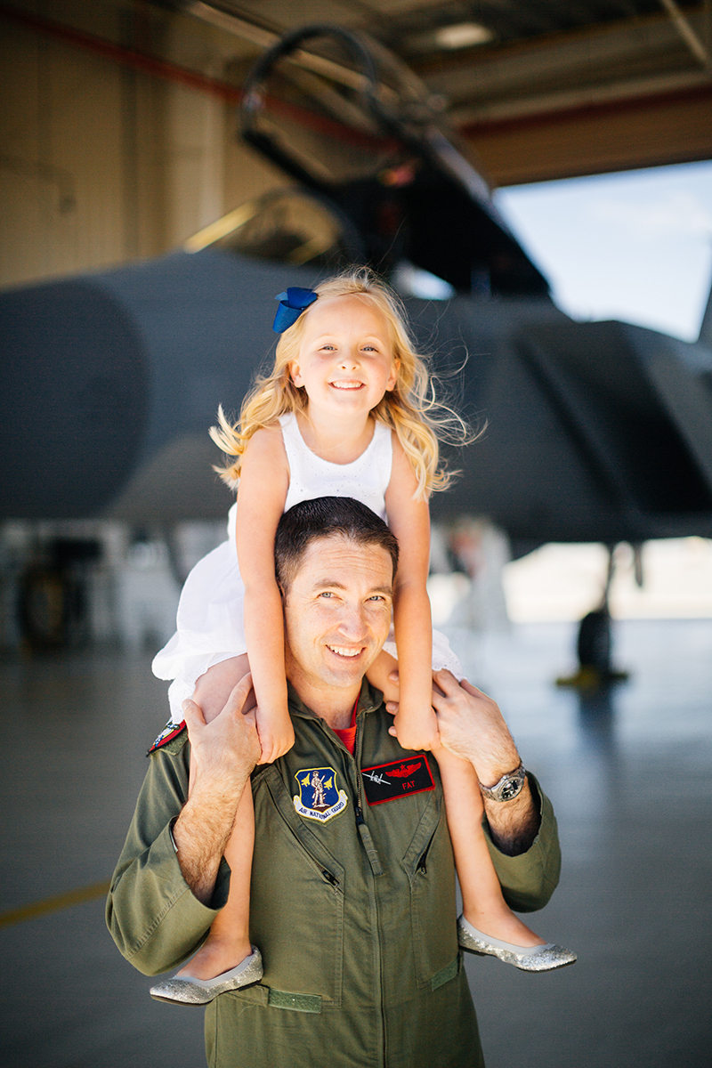 A pilot holds his daughter on his shoulders in front of an F-15 at Barnes Air National Guard Base wearing a flight suit and a coordinated outfit for these F-15 fighter pilot family photos