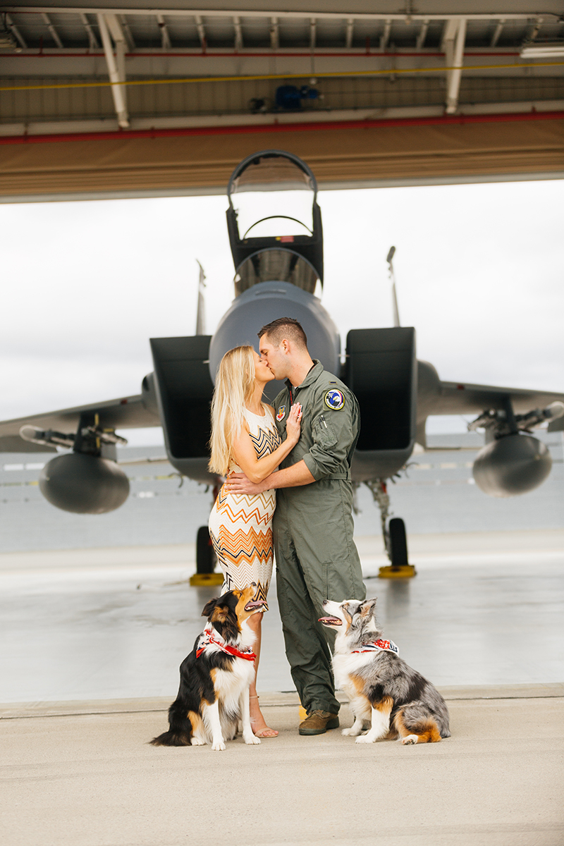 A pilot and his wife stand together kissing with their dogs in front an F-15 at Barnes Air National Guard Base wearing a flight suit and coordinated outfits for these F-15 fighter pilot family photos