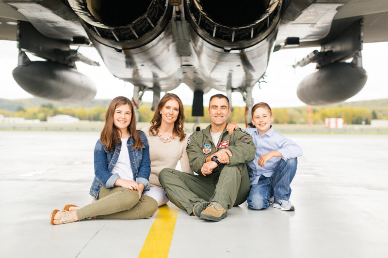 A family sits under an F-15 holding each other close at Barnes Air National Guard Base wearing a flight suit and coordinated outfits for these F-15 fighter pilot family photos