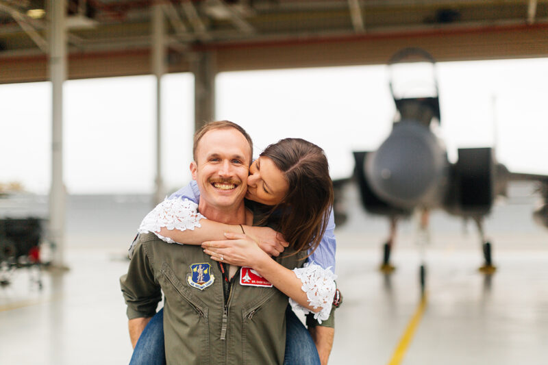 A pilot gives his wife a piggyback ride in front of an F-15 at Barnes Air National Guard Base wearing a flight suit and a coordinated outfit for these F-15 fighter pilot family photos