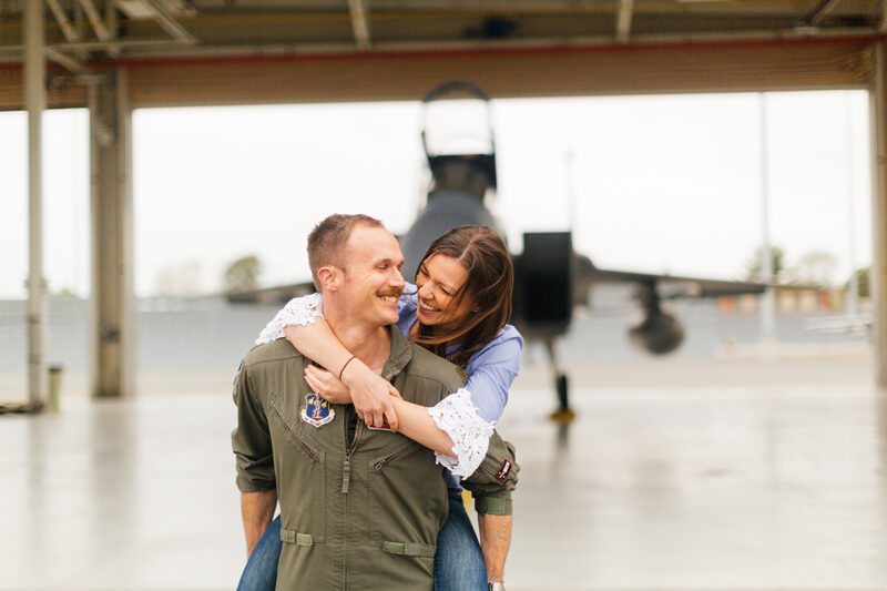 A pilot gives his wife a piggyback ride in front of an F-15 at Barnes Air National Guard Base wearing a flight suit and a coordinated outfit for these F-15 fighter pilot family photos