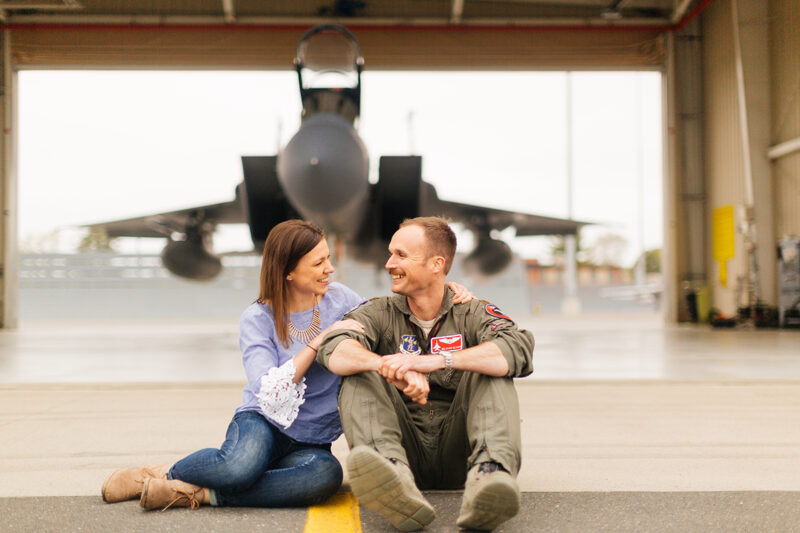 A pilot and his wife sit together in front of an F-15 at Barnes Air National Guard Base wearing a flight suit and a coordinated outfit for these F-15 fighter pilot family photos