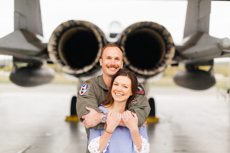 A pilot and his wife stand together behind an F-15 at Barnes Air National Guard Base wearing a flight suit and a coordinated outfit for these F-15 fighter pilot family photos