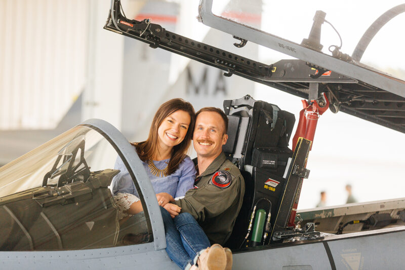 A pilot sits in the cockpit of an F-15 with his wife sitting in his lap at Barnes Air National Guard Base wearing a flight suit and a coordinated outfit for these F-15 fighter pilot family photosA pilot sits in the cockpit of an F-15 fighter with his wife sits in his lap in a hangar at Barnes Air National Guard Base near Boston wearing a flight suit and a coordinated outfit for these F-15 fighter pilot family photos