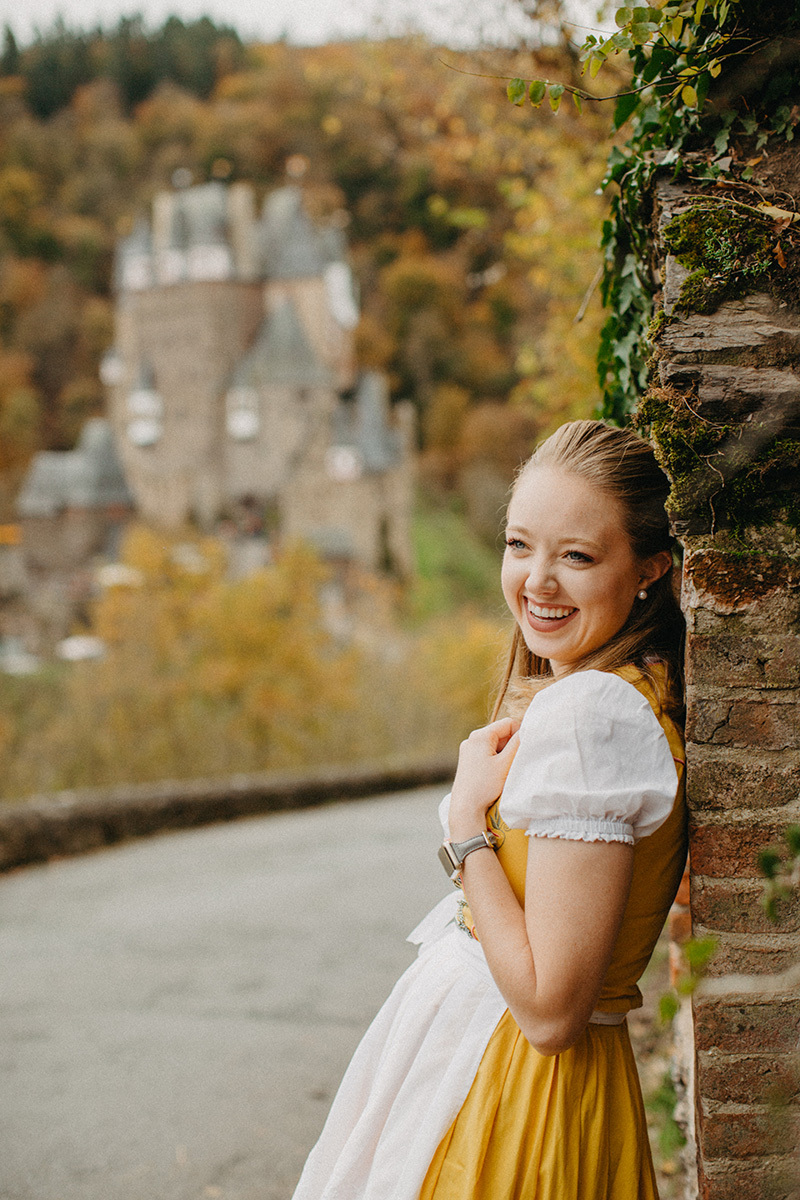 A beautiful young blonde woman poses on the road to Burg Eltz wearing a traditional dirndl for these Eltz Castle couples photos in Germany